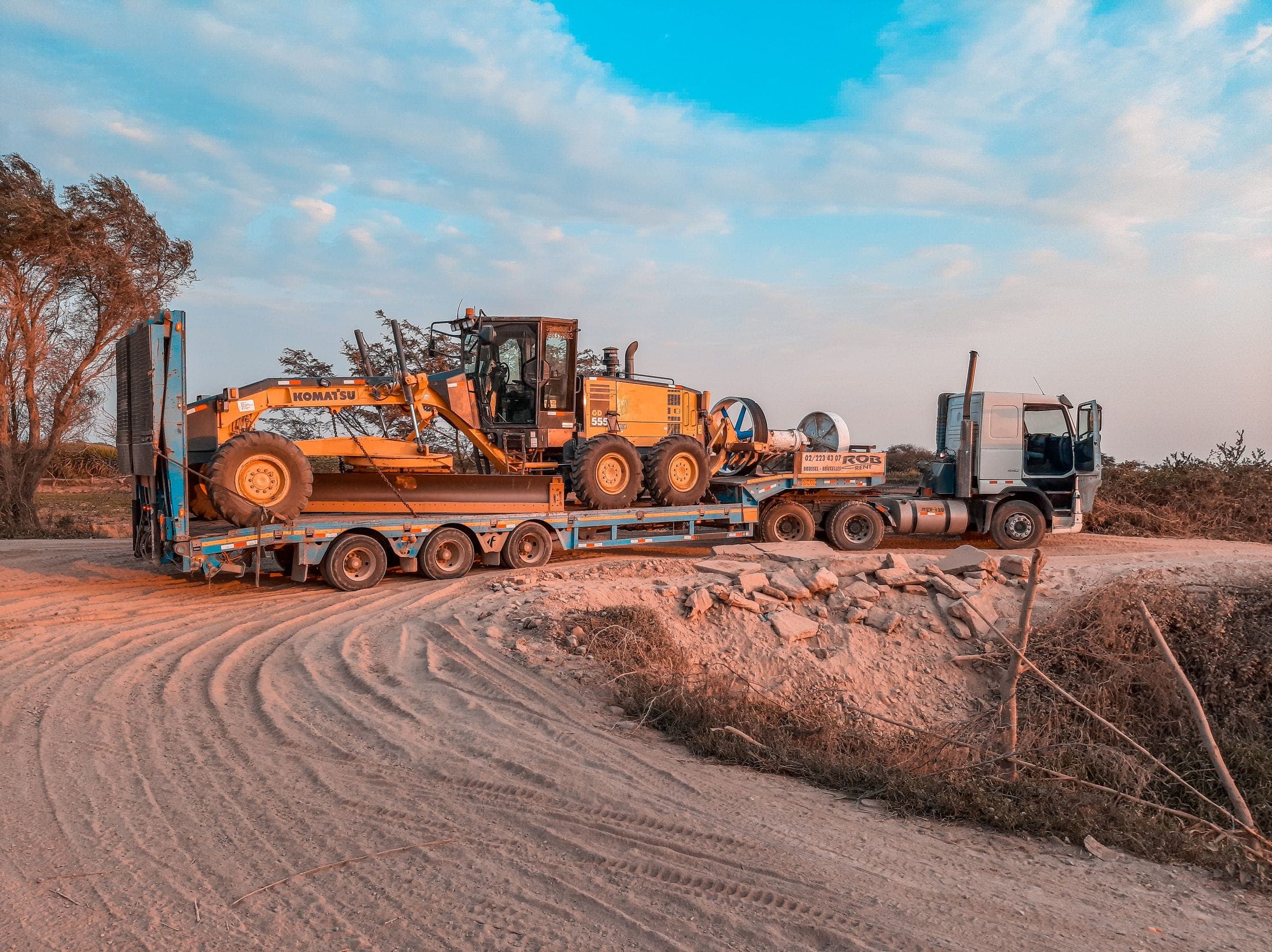 A low-bed trailer carrying a motor grader on a dirt road